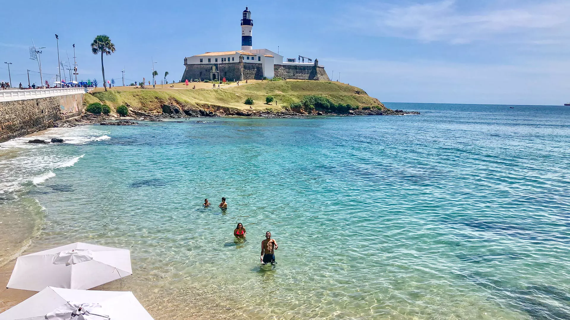 Pessoas na água pouco profunda próximo a um farol histórico à beira-mar em uma cidade litorânea.
